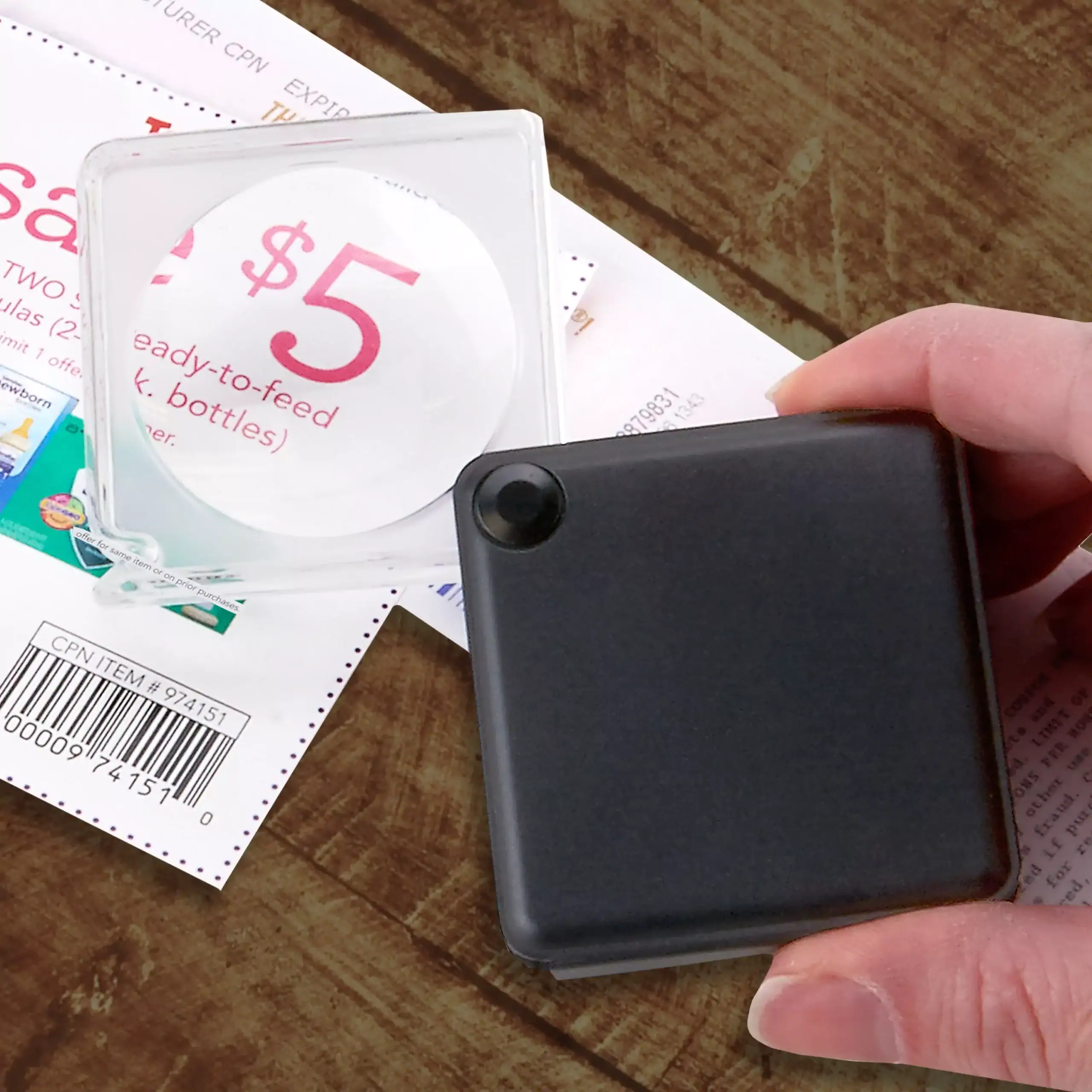 Una mano sostiene la lupa de bolsillo abatible Carson GN-33 MagniFlip™ 3x de 3,8 cm sobre un cupón, ampliando el texto y código de barras sobre una superficie de madera.