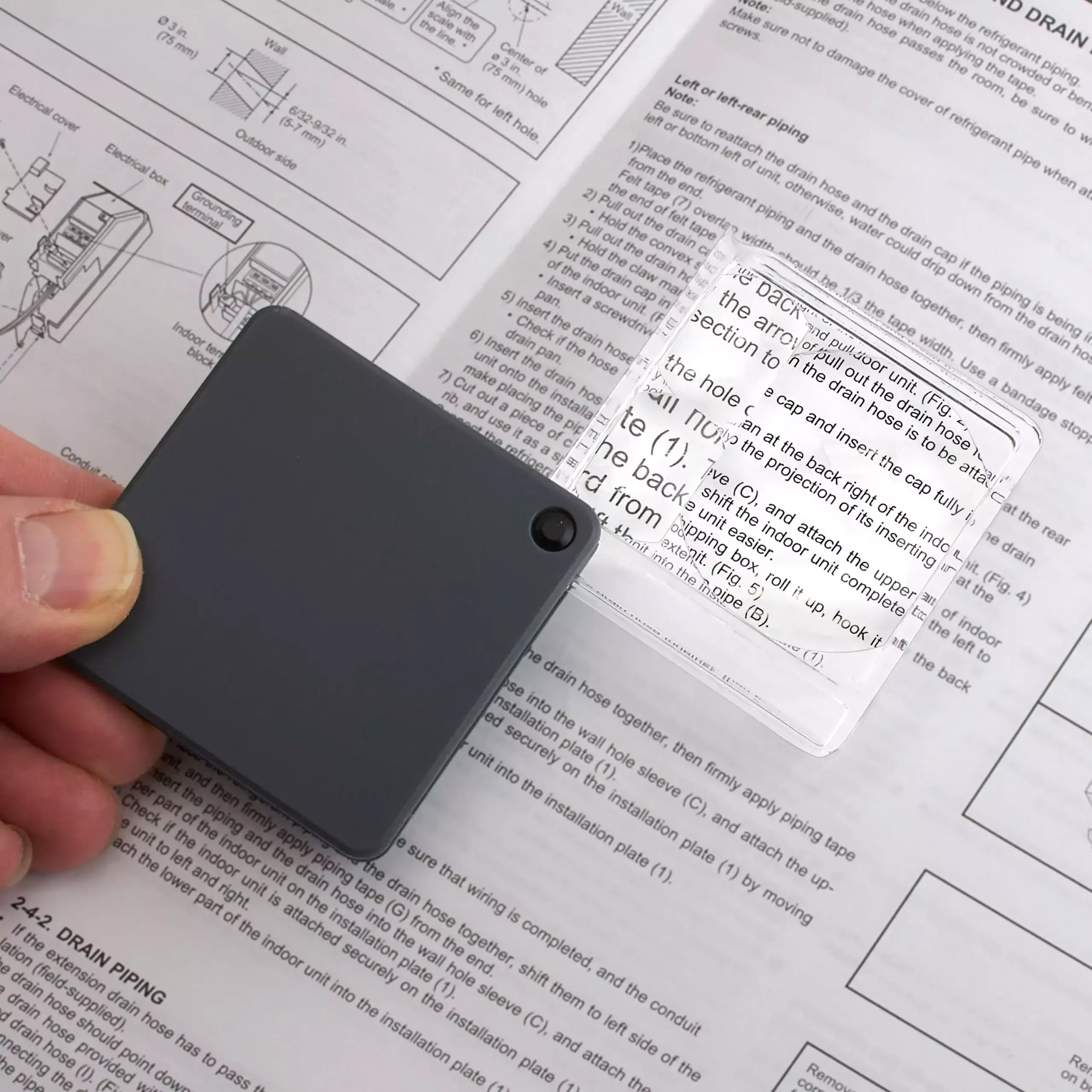 Una persona sostiene una lupa de bolsillo abatible Carson GN-44 Serie MagniFlip™ Plus 2.5x/5x/7x con funda protectora y lentes de cristal sobre un manual impreso, ampliando el texto que está debajo de la lente.