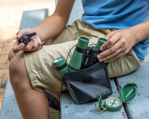 Una persona se sienta en un banco con una brújula y el Kit infantil de exploración 5 en 1 Carson HU-401 | Serie AdventurePak™, teniendo prismáticos y linterna del kit guardados en su mochila para aventuras STEM.