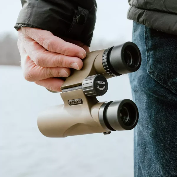 Una persona sostiene los prismáticos compactos Carson HW-025 Serie Stinger™ 10x25 mm con óptica avanzada y diseño ligero junto al agua, usando chaqueta oscura y jeans: ideales para disfrutar de la naturaleza en cualquier ocasión.