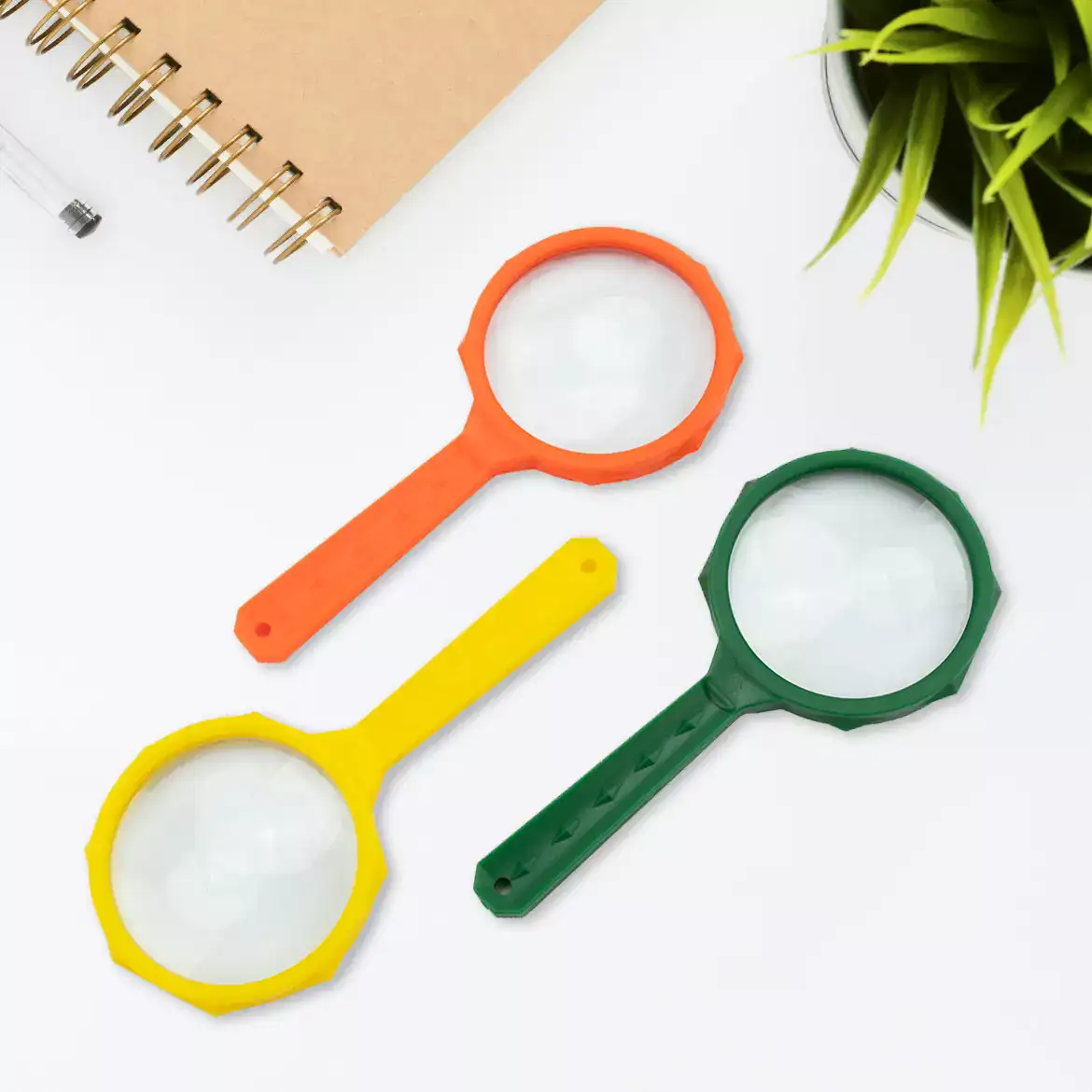 Cincuenta lupas infantiles de cristal Carson JD-3MU MagniRama™ 3x, cada una con una lente de 50 mm y mangos de colores surtidos, se exhiben sobre una superficie blanca cerca de un cuaderno, un bolígrafo y una planta verde en maceta.
