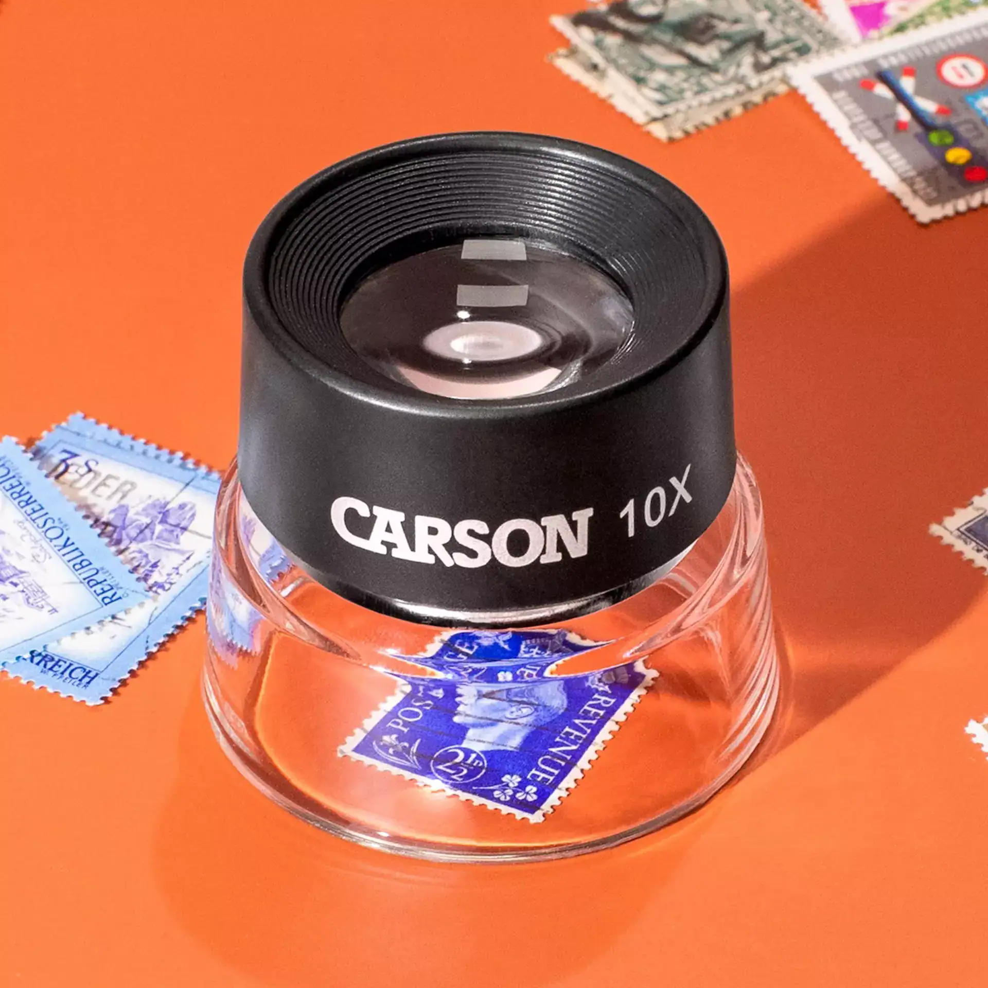 La lupa de pie tipo cuentahilos Carson LL-10 Serie LumiLoupe® 10x con doble lente y base de 1" descansa sobre un sello postal azul, rodeada de otros sellos esparcidos en una superficie naranja.