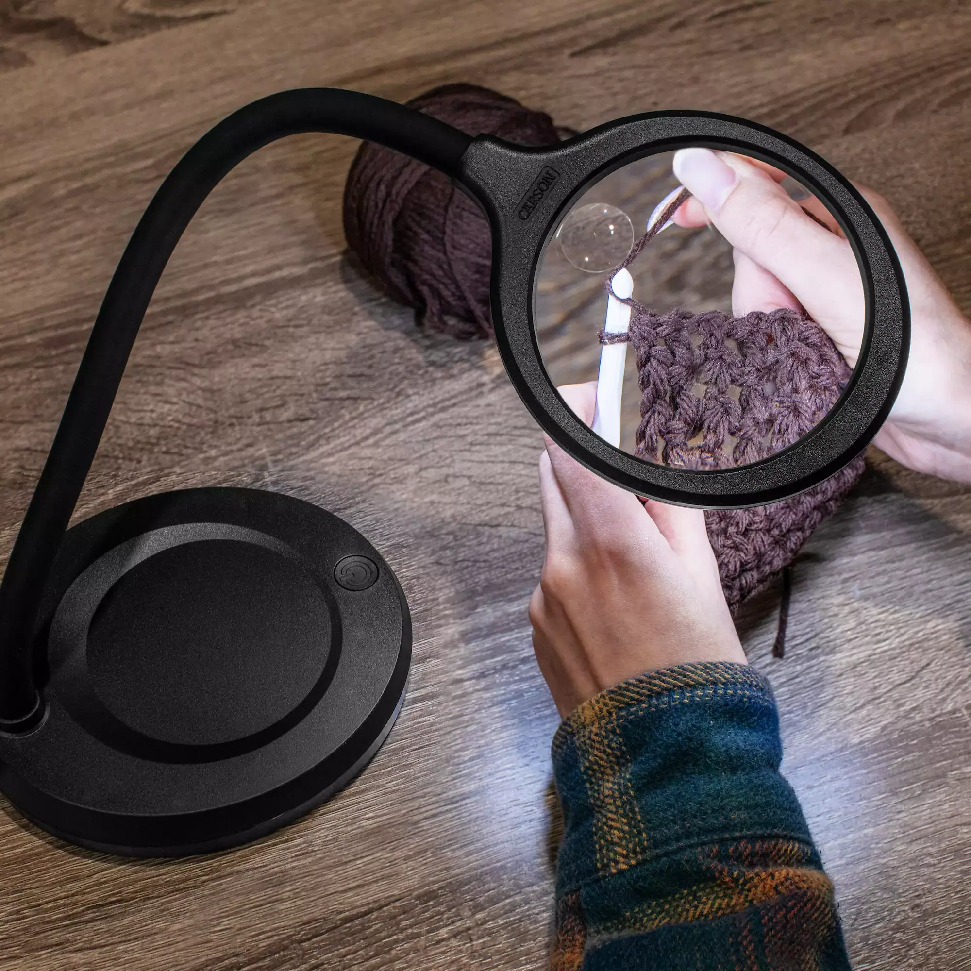 Una persona usa la lámpara lupa de mesa con LED COB Carson LM-30 Serie DeskBrite™ 2x, con lente asférica de 4" y cuello flexible, para tejer con hilo marrón y ganchillo sobre una superficie de madera.
