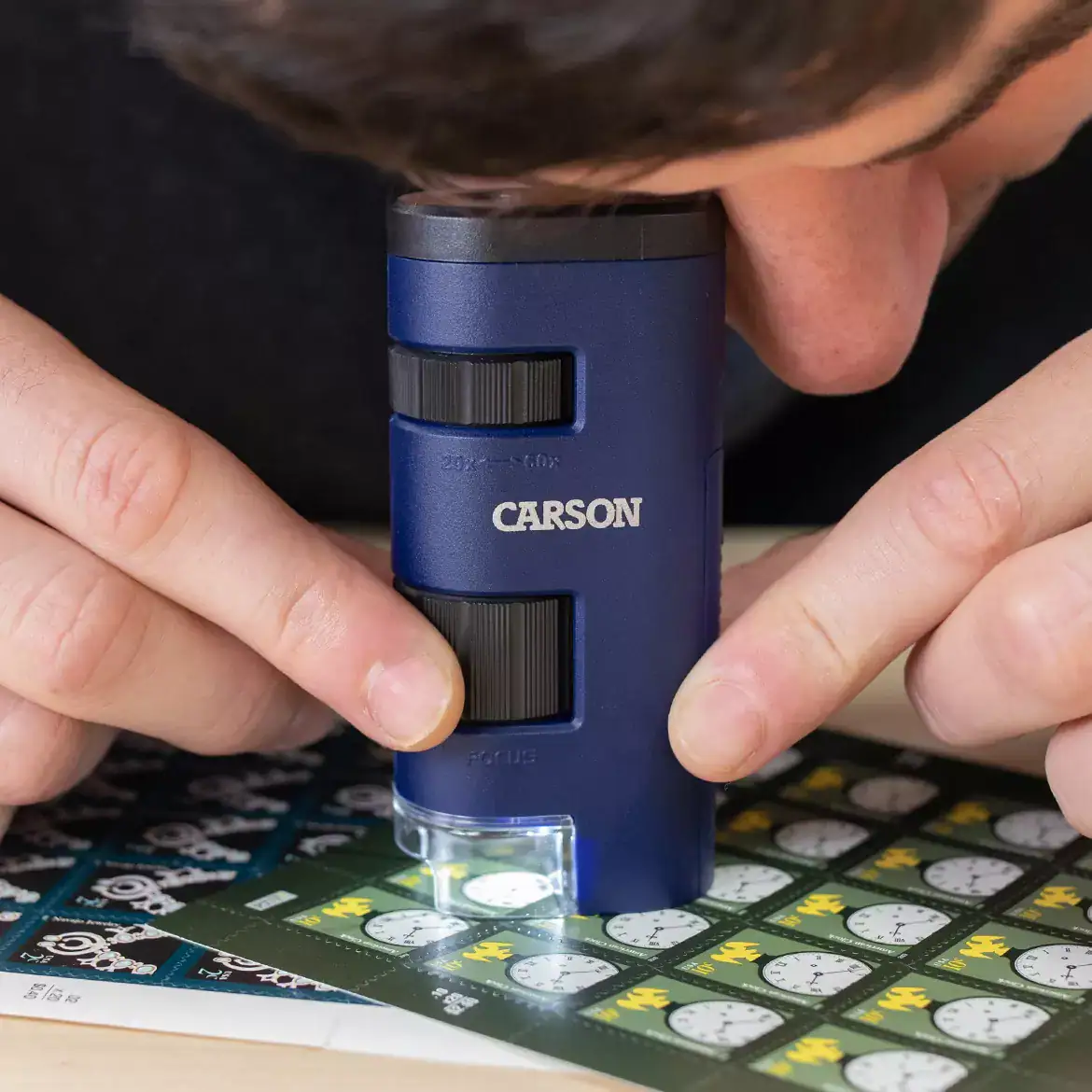 Una persona usa el Microscopio de bolsillo Carson MM-450 | Serie Pocket Micro™ 20x-60x LED portátil para observar detalles en pegatinas de relojes animados, despertando su curiosidad y pasión por la exploración STEM.