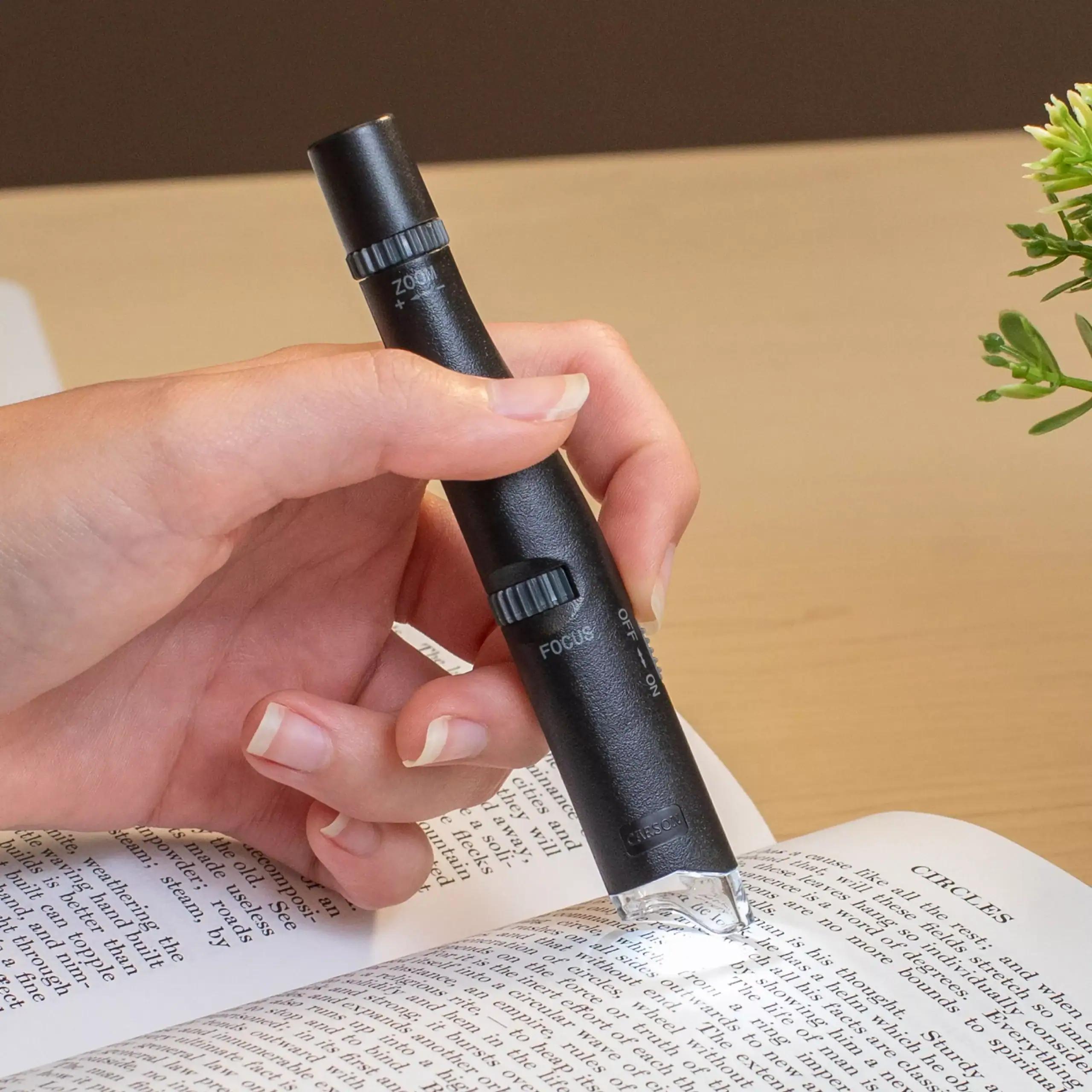 Una mano sostiene el Microscopio de bolsillo Carson MP-300 | Serie MicroPen™ 24x-53x con luz LED sobre un libro abierto, ampliando el texto para facilitar la lectura.