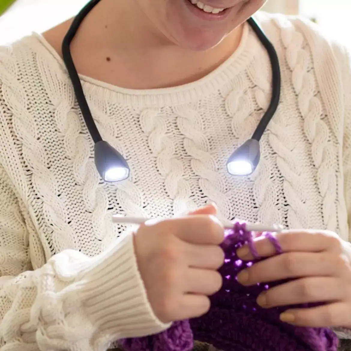 Una persona con un suéter blanco teje con estambre morado mientras usa la Luz de cuello LED COB Carson NL-10, que tiene doble cabezal, brillo ajustable, diseño manos libres y cuello flexible.