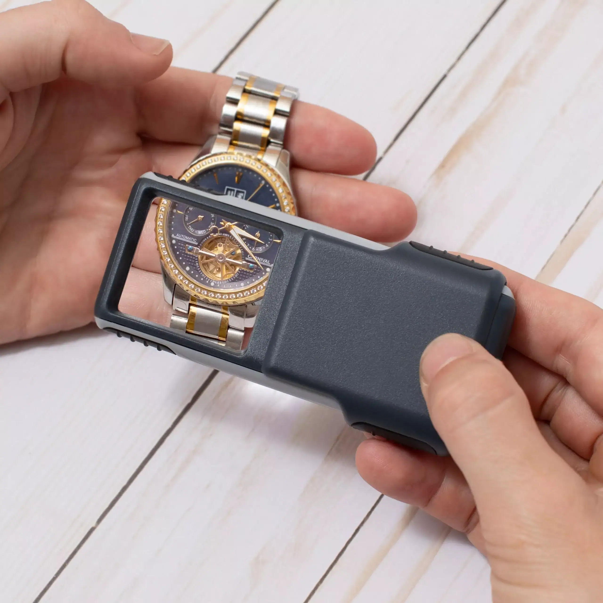 Una persona utiliza la lupa portátil deslizante con LED Carson PO-25 de la serie MiniBrite™ 3x para inspeccionar un reloj de pulsera con correa metálica y esfera azul sobre una superficie de madera blanca.