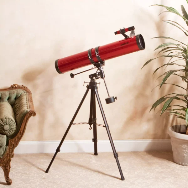 Un Telescopio reflector Carson RP-300 Serie Red Planet™ 45-100x114 mm con montura ecuatorial y óptica de precisión se encuentra sobre un trípode negro junto a una butaca verde ornamentada y una planta en maceta en una habitación alfombrada.