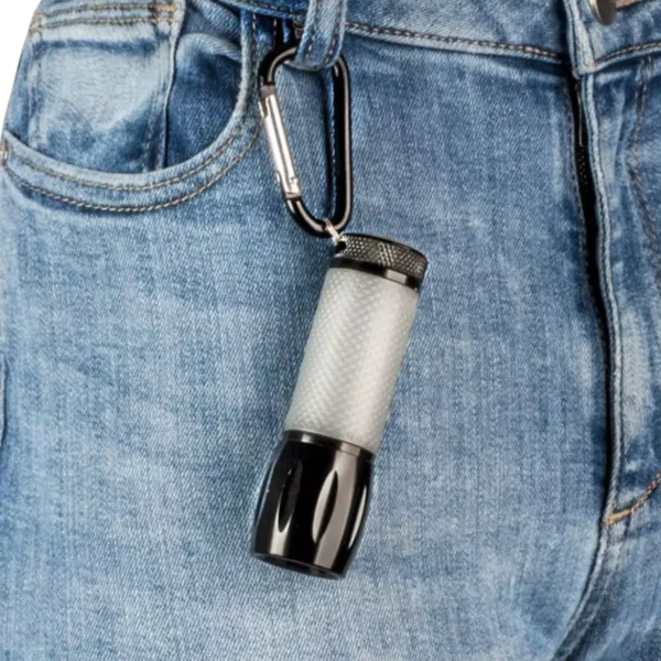 La Mini linterna LED Carson SL-55 | Serie LEDSight Pro™ con mango que brilla en la oscuridad y diseño compacto está sujeta al cinturón de unos jeans de mezclilla azules con un clip tipo mosquetón.