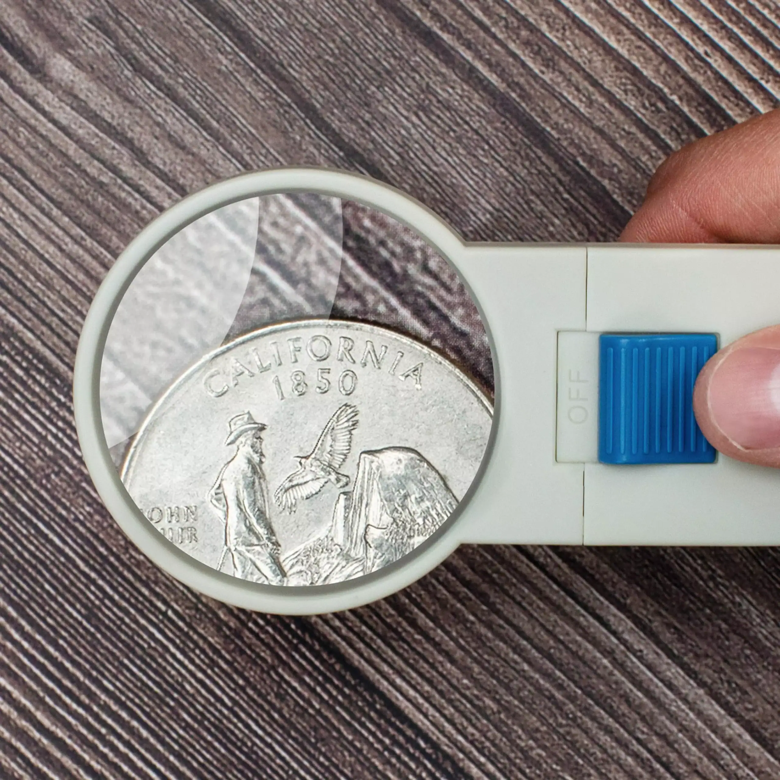Una mano sostiene la lupa de mano con LED Carson SV-50 HandHeld™ 5x con lente asférica de 2,5" sobre una moneda de California, ampliando sus detalles en una superficie de madera.