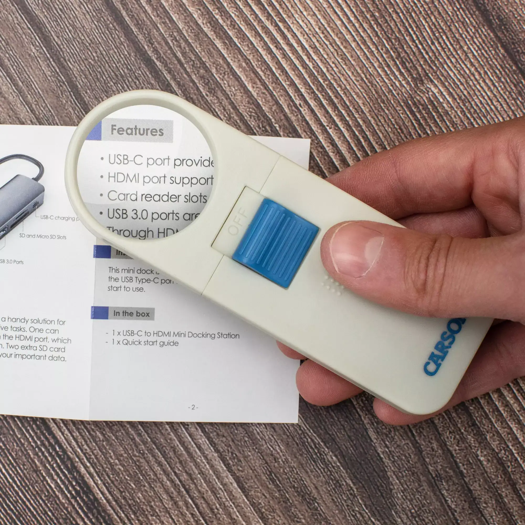 Una mano sostiene una lupa de mano compacta Carson SV-70 HandHeld™ 7x de 1,5" (3,8 cm) rectangular con lente asférica y interruptor azul sobre un manual impreso en una superficie de madera.