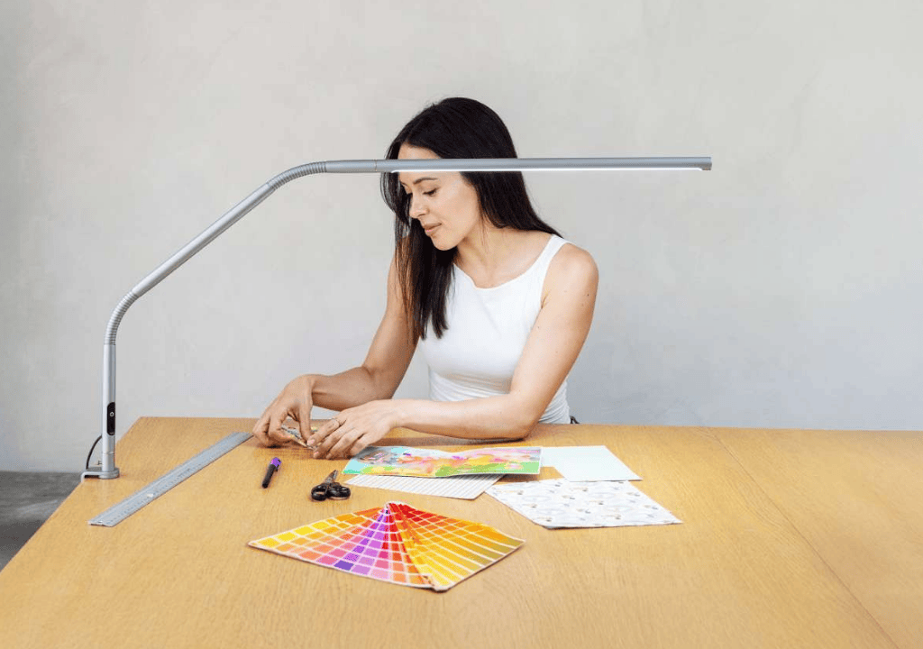Una mujer se sienta en una mesa de madera bajo una lámpara de escritorio, trabajando con papeles de colores y materiales de diseño, incluyendo una muestra de color y una regla.
