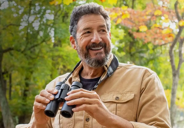 Hombre con barba y camisa canela sostiene unos prismáticos al aire libre, rodeado de árboles verdes y otoñales.